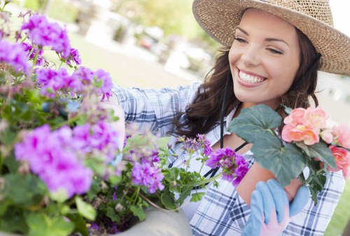 A woman with long dark hair, wearing a wide-brimmed straw hat and light blue gardening gloves, is tending to a colorful flower bed in a well-maintained suburban garden. The flower bed includes vibrant purple, pink, and peach blossoms, surrounded by lush green foliage. The garden features a neatly trimmed lawn with densely planted hedges and small trees in the background, illuminated by bright, natural daylight suggesting a clear, sunny day. The scene captures the outdoor environment of a typical back garden in Acton, with a focus on the arrangement of flowering plants alongside paved pathways or patio areas. The woman is smiling as she carefully arranges the flowers, indicating a moment of gardening activity supported by professional gardening services such as those offered by Garden Maintenance Acton. The overall setting showcases a thoughtfully landscaped garden environment with attention to plant health, layout, and natural tones, reflecting the quality of outdoor maintenance and gardening expertise.