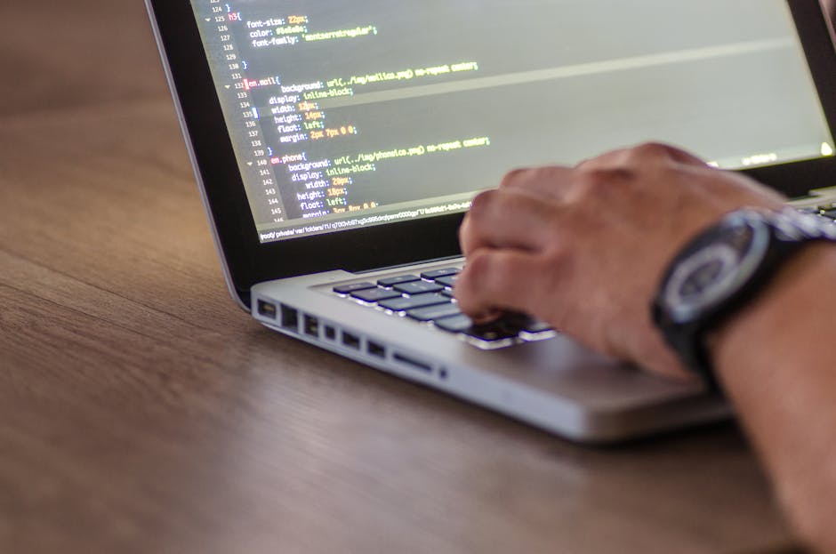 A person with a wristwatch is using a laptop placed on a wooden surface, typing on the keyboard. The laptop screen displays lines of code with a dark background and highlighted syntax. The scene sugge