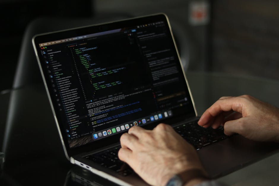 A person with a wristwatch is using a laptop placed on a wooden surface, typing on the keyboard. The laptop screen displays lines of code with a dark background and highlighted syntax. The scene sugge